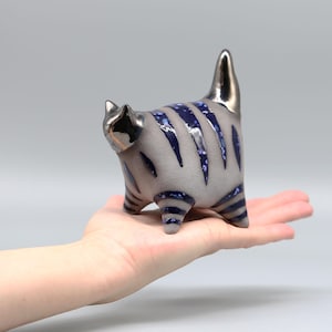 May include: A ceramic cat figurine with a gray and blue striped pattern. The cat is standing on its hind legs with its tail raised in the air.