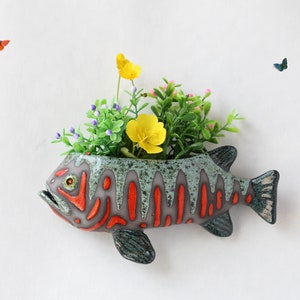 Op de afbeelding: Een keramische vis-plantenpot met een groene, oranje en witte glazuur. De pot is gevuld met groene planten en gele bloemen. Twee vlinders vliegen in de achtergrond.