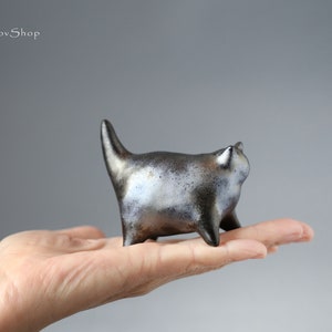 Könnte beinhalten: Eine Keramik-Katzenfigur mit schwarzer und silberner Glasur. Die Katze steht auf allen vieren und hat ihren Schwanz eingerollt. Die Figur ist etwa 10 cm hoch.