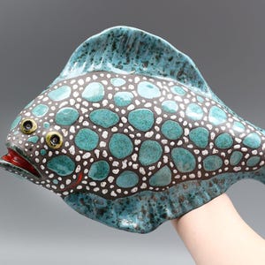 Könnte beinhalten: Eine Keramikfischskulptur mit einem türkisfarbenen und braunen gepunkteten Muster. Der Fisch hat einen roten Mund, gelbe Augen und eine türkisfarbene Flosse. Die Skulptur wird von einer Hand gehalten.