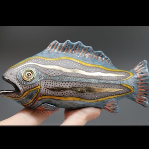 May include: A ceramic fish sculpture featuring a blue body with a textured, dotted grey midsection. The fish is decorated with horizontal yellow and gold stripes, a prominent eye, and orange-tipped fins. The mouth is open, showing a dark interior.