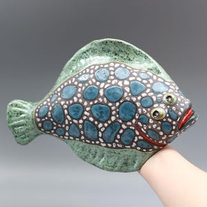 Op de afbeelding: Een keramische vissculptuur met een groene body en staart. De vis heeft een patroon van blauwe en witte stippen op zijn lichaam, met een donkerbruine omtrek. De vis heeft rode lippen en kleine, gedetailleerde ogen.