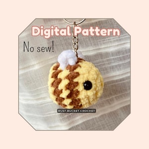 May include: A crocheted bee keychain with brown and yellow stripes, white fuzzy wings, and black eyes. The keychain is attached to a silver key ring. The text "Digital Pattern No sew!" is visible in the image.