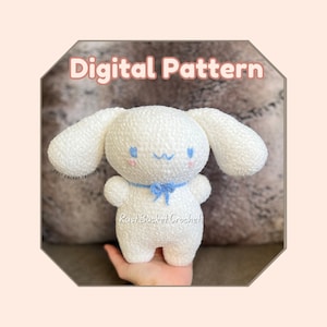 May include: A white crocheted plush toy with blue eyes and a blue bow. The toy is shaped like a dog with floppy ears and a small, round body. The toy is being held in a hand. The text "Digital Pattern" is at the top of the image. The text "RustBucketCrochet" is at the bottom of the image.