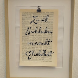 Könnte beinhalten: Ein gerahmter Druck mit dem deutschen Text "Zu viel Nachdenken verursacht Grübelei" wird über einem offenen Buch angezeigt. Zwei Kerzenhalter mit brennenden Kerzen flankieren den Rahmen. Das Buch hat gealterte Seiten und einen dekorativen Einband.