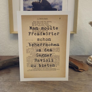 Könnte beinhalten: Ein gerahmter Druck mit deutschem Text, darunter die Worte "Man sollte Fremdwörter schon beherrschen" in einer Vintage-Schriftart. Der Druck befindet sich auf einer Holzoberfläche, mit einem Gemälde im Hintergrund.