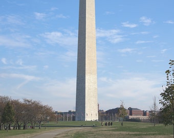 May include: A tall, white marble obelisk, the Washington Monument, stands against a clear blue sky with white clouds. A paved path leads to the monument, with green grass and trees in the foreground.