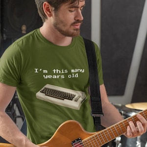 May include: A man wearing a green t-shirt with the text "I'm this many years old" and a retro computer keyboard graphic. He is playing a brown electric guitar.
