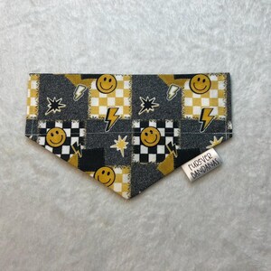 Può includere: Una bandana triangolare per animali domestici con un motivo patchwork di quadrati a scacchi bianchi e neri, quadrati bianchi e gialli, faccine sorridenti gialle e fulmini neri. Un'etichetta in tessuto bianco riporta FUREVER BANDANAS.