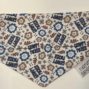 Könnte beinhalten: Ein dreieckiges Haustierhalstuch mit weißem Hintergrund, das ein wiederkehrendes Muster aus blauen und braunen Blumen, kleinen Blättern und dem Text "GRUMPY FUCKER" in dunkelblauen Buchstaben zeigt. Ein kleines weißes Etikett trägt "FUREVER BANDANAS".