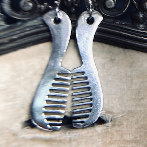 May include: A pair of silver earrings with a comb design. The earrings are hanging from a black metal stand.