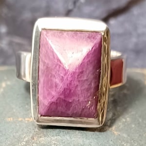 May include: A silver ring with a square-shaped, purple gemstone set in a silver bezel. The ring has a Celtic knot design around the bezel.