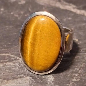 May include: A silver ring featuring an oval tiger's eye gemstone. The gemstone is a rich golden brown colour with a distinctive chatoyancy effect.