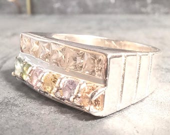 May include: A silver-toned ring featuring two horizontal rows of gemstones. The top row contains square-cut champagne-colored stones, while the bottom row displays a series of round-cut multicolored stones in shades of yellow, pink, and green.