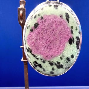 Pu&ograve; includere: Ciondolo ovale con castone color argento. Il ciondolo presenta un'area rosa centrale circondata da una base verde chiaro con macchie nere. &Egrave; sospeso da un'ansa color argento.