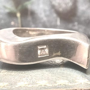 May include: A silver ring with a unique, curved design. The ring features a square, clear gemstone set in the center. The metal has a polished finish, reflecting light. The ring is a piece of jewelry.