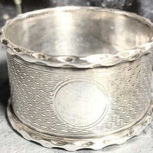 May include: A silver napkin ring with a wavy pattern and a circular, blank space. The top and bottom edges have a scalloped design. The ring is made of a shiny, reflective metal.