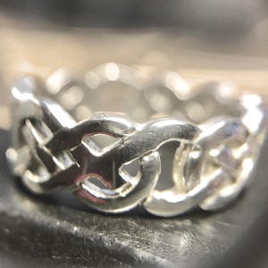 May include: A silver-colored ring with a woven chain design. The ring has a continuous pattern of interlocking links, creating a textured surface. The metal reflects light, highlighting the intricate details of the design.