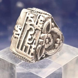Op de afbeelding: Een zilverkleurige ring met ingewikkelde bloemen- en gestileerde tekstdetails. De ring heeft een rechthoekige voorkant met gegraveerde karakters en een bloemenontwerp aan de zijkant. De ring wordt gepresenteerd op een helder, schuin oppervlak.