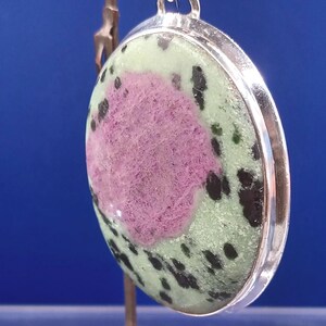 Pu&ograve; includere: Ciondolo rotondo con castone color argento. La pietra presenta un'area rosa centrale circondata da verde e macchie nere. Il ciondolo &egrave; esposto su un piccolo supporto su sfondo blu.
