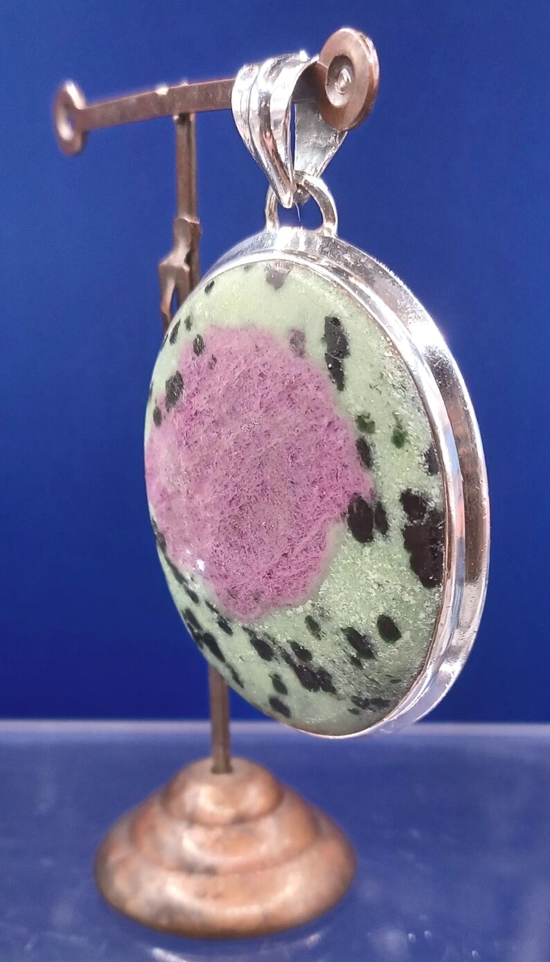 Pu&ograve; includere: Ciondolo rotondo con castone color argento. La pietra presenta un'area rosa centrale circondata da verde e macchie nere. Il ciondolo &egrave; esposto su un piccolo supporto su sfondo blu.