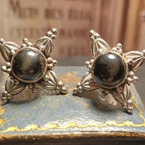 May include: A pair of antique silver-toned earrings. Each earring features a large, dark, round stone at the center, surrounded by a decorative, starburst design. The earrings are displayed on a vintage blue box.