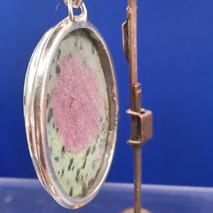 Pu&ograve; includere: Ciondolo ovale con cornice color argento, con una pietra rosa e verde. Il ciondolo &egrave; sospeso a un anello d'argento. &Egrave; esposto su un supporto color rame su sfondo blu.