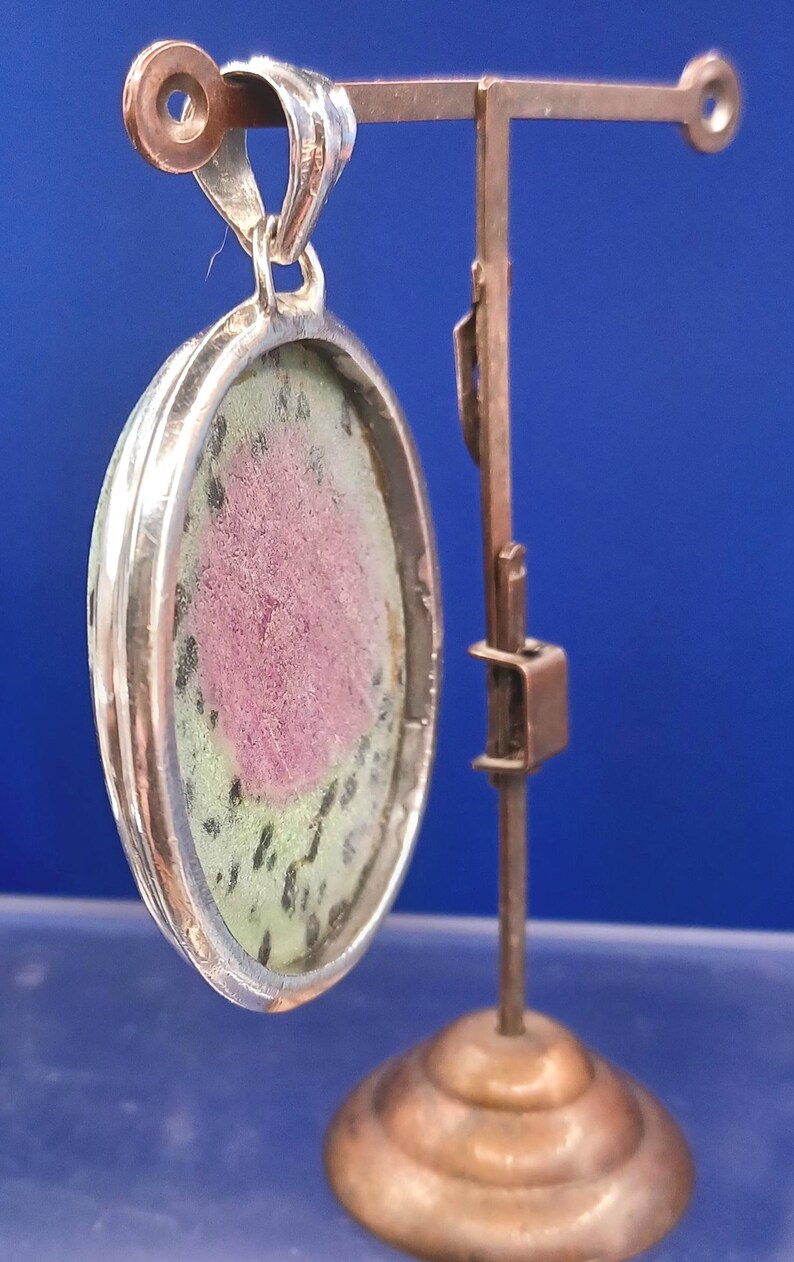 Pu&ograve; includere: Ciondolo ovale con cornice color argento, con una pietra rosa e verde. Il ciondolo &egrave; sospeso a un anello d'argento. &Egrave; esposto su un supporto color rame su sfondo blu.