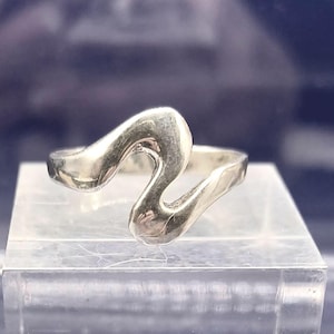 Puede incluir: Un anillo plateado con un diseño abstracto y fluido. El anillo presenta una forma ondulada y curvada que se envuelve alrededor del dedo. El anillo se exhibe sobre una superficie clara y rectangular sobre un fondo oscuro.