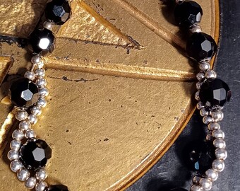 May include: A black beaded bracelet with silver accents. The bracelet is made of faceted black beads and small silver beads. The bracelet is on a gold background.