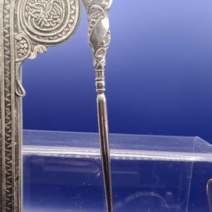 May include: A silver letter opener with an ornate handle and a decorative stand. The stand features Celtic knotwork and a stylized animal design. The opener is displayed against a blue background.