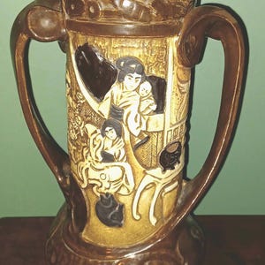 May include: Brown ceramic vase with two handles, featuring a relief design of a family scene. The vase has a wide base and a detailed, textured surface. The color palette includes shades of brown, beige, and black.