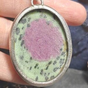 Pu&ograve; includere: Ciondolo ovale con cornice color argento. Il ciondolo presenta una pietra rosa centrale circondata da una pietra verde e nera maculata. Il ciondolo &egrave; sospeso a una catena color argento.