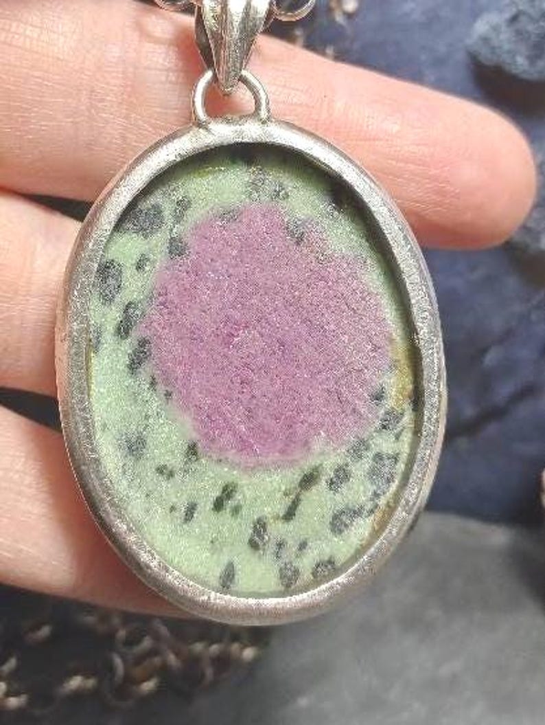 Pu&ograve; includere: Ciondolo ovale con cornice color argento. Il ciondolo presenta una pietra rosa centrale circondata da una pietra verde e nera maculata. Il ciondolo &egrave; sospeso a una catena color argento.