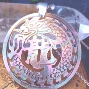 May include: A circular pendant with a dragon design, made from iridescent mother-of-pearl. The pendant has a silver bail and hangs from a fine chain. The dragon is intricately cut out, revealing the background. The pendant has a pearlescent sheen.
