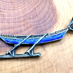 May include: A silver-toned metal pendant shaped like a long, narrow boat with two oars extending downward. The boat features a vibrant blue enamel inlay along its hull and includes a circular jump ring at the top for attachment to a chain or cord.