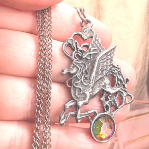 Può includere: Una collana con ciondolo in metallo color argento raffigurante un cavallo alato dal design traforato. Il ciondolo è appeso a una catenina color argento abbinata e presenta una pietra rotonda sfaccettata iridescente e multicolore alla base.