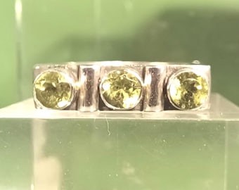 May include: A silver ring with three round, light green gemstones. The gemstones are set in a rectangular band with vertical silver bars separating each stone. The ring is displayed on a clear, angled surface.