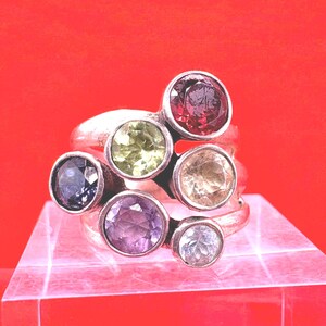 May include: A silver ring featuring seven colorful gemstones in various sizes. The gemstones include red, green, purple, blue, and clear stones. The ring is displayed on a clear, angled surface against a red background.