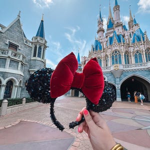 Puede incluir: Diadema de orejas de Minnie Mouse de lentejuelas negras y rojas con un gran lazo rojo. La diadema se sostiene frente a un gran castillo rosa y azul.