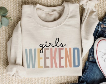 May include: A cream colored sweatshirt with the text "girls weekend" in colorful letters. The sweatshirt is laying on a beige carpet with a pair of blue jeans and white sneakers.