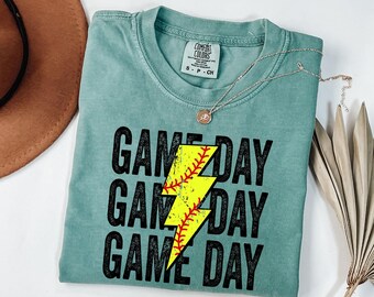 May include: A teal Comfort Colors t-shirt with the words "GAME DAY" stacked three times. A yellow lightning bolt graphic with a baseball design is in the center. A silver necklace with a small pendant is draped across the shirt.
