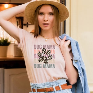 May include: A woman wearing a light pink t-shirt with a leopard print paw print graphic and the text "DOG MAMA" repeated in different colors. She is wearing a straw hat and a denim jacket.