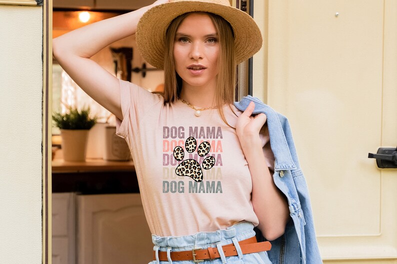 May include: A woman wearing a light pink t-shirt with a leopard print paw print graphic and the text "DOG MAMA" repeated in different colors. She is wearing a straw hat and a denim jacket.