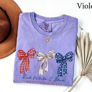 May include: A violet short-sleeved t-shirt featuring three bows in red gingham, white with gold trim, and blue with white stars. Below the bows, the text reads Red White & Blue America 250 Years. The shirt has a Comfort Colors brand neck label.