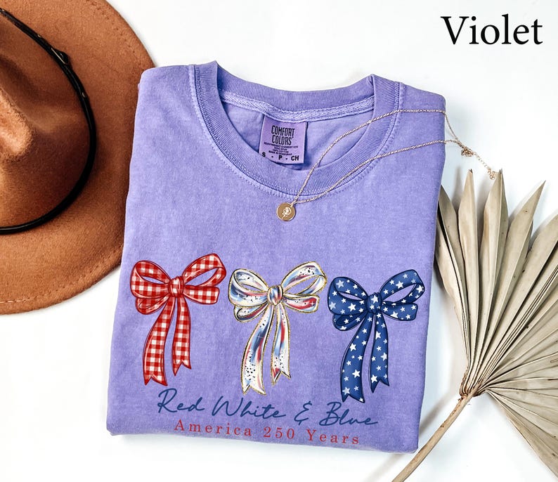 May include: A violet short-sleeved t-shirt featuring three bows in red gingham, white with gold trim, and blue with white stars. Below the bows, the text reads Red White & Blue America 250 Years. The shirt has a Comfort Colors brand neck label.