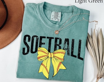 May include: Light green t-shirt with the word "SOFTBALL" in black block letters and a yellow softball bow graphic. The shirt has a crew neck and short sleeves. A silver necklace with a pendant is draped across the shirt.
