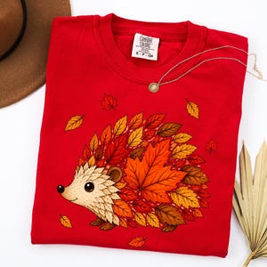 May include: A bright red t-shirt featuring a graphic of a hedgehog whose body is composed of various autumn-colored leaves in shades of orange, yellow, and brown. The shirt has a crew neckline and a white label reading Comfort Colors S-P-CH.