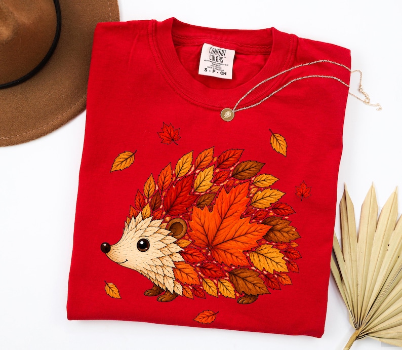 May include: A bright red t-shirt featuring a graphic of a hedgehog whose body is composed of various autumn-colored leaves in shades of orange, yellow, and brown. The shirt has a crew neckline and a white label reading Comfort Colors S-P-CH.