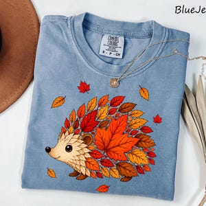 May include: A blue jean colored t-shirt featuring a graphic of a hedgehog whose back is covered in vibrant autumn leaves in shades of red, orange, and brown. The shirt has a crew neckline and a Comfort Colors brand label on the inside collar.